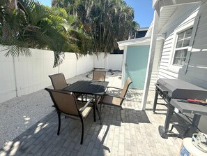 A patio with a table and chairs, a grill, and a white fence with palm trees. - Cozy Beach Cottage, Pet Friendly (Holmes Beach)