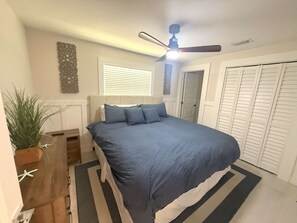 A bedroom with a bed, a ceiling fan, a window with blinds, a potted plant, a wooden nightstand, and a closet with sliding doors. - Cozy Beach Cottage, Pet Friendly (Holmes Beach)