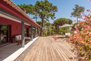 Een houten terras met buitenmeubilair, een rood gebouw en omliggende bomen.