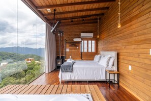 A wooden cabin bedroom with a large bed, a small table, and a view of the mountains. - Apt w/ Fireplace, Jacuzzi, and Amazing View Vst027 (Anitapolis)