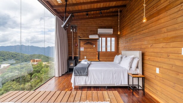 A wooden cabin bedroom with a large bed, a small table, and a view of the mountains. - Apt w/ Fireplace, Jacuzzi, and Amazing View Vst027 (Anitapolis)