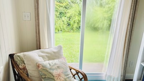 A room with a wooden-framed sofa bed with floral upholstery, a view of a green garden through a large window, and a blue wooden floor.