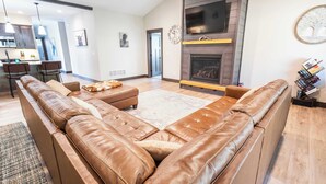 Ein modernes Wohnzimmer mit einem großen Ledersofa, einem Kamin, einem Flachbildfernseher und einer Küche im Hintergrund.