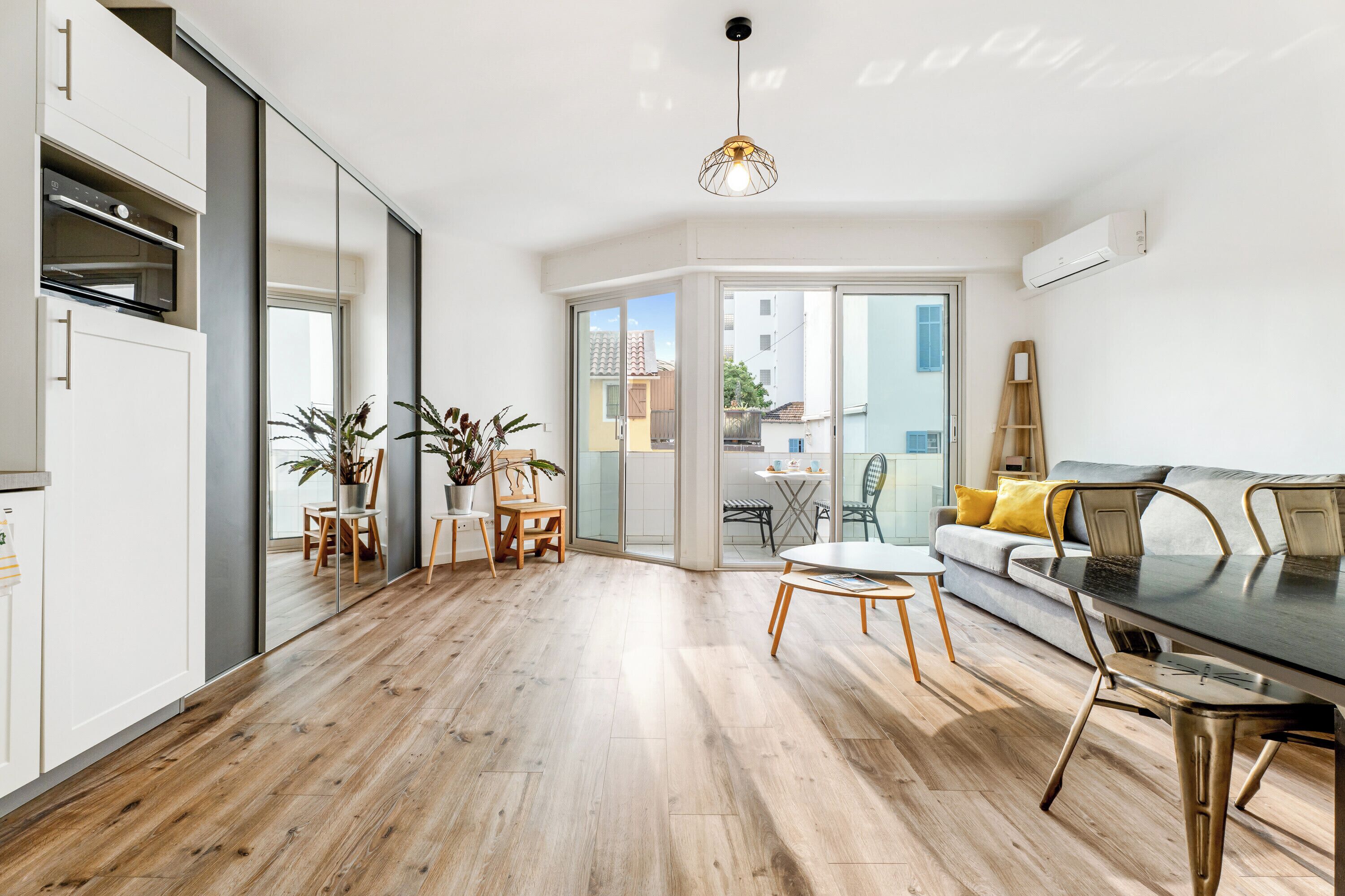 A modern apartment with a kitchen, dining area, and living room. It features wooden flooring, a dining table with chairs, a sofa, and a large window providing a view of the outside.