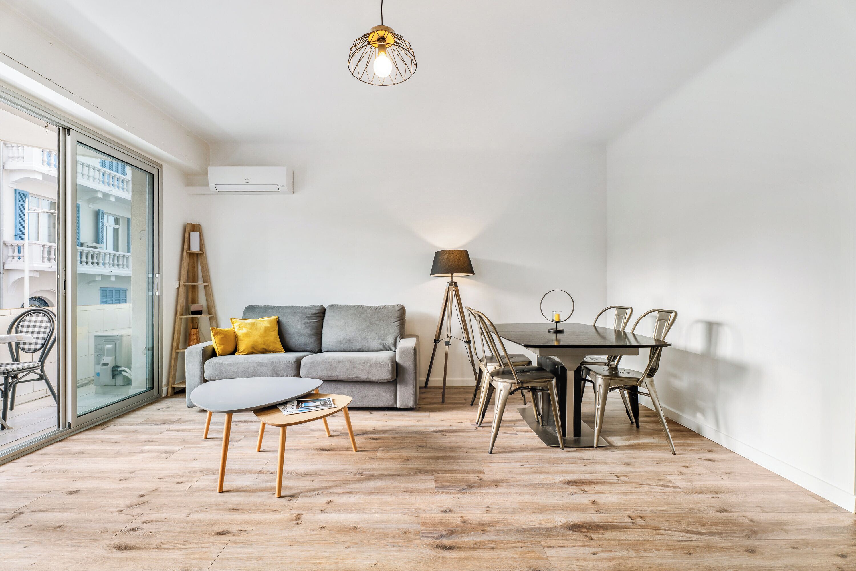 Uma sala de estar moderna com um sofá cinzento, mesa de centro de madeira e uma mesa de jantar com cadeiras. Há uma janela grande que oferece luz natural e vista para edifícios.
