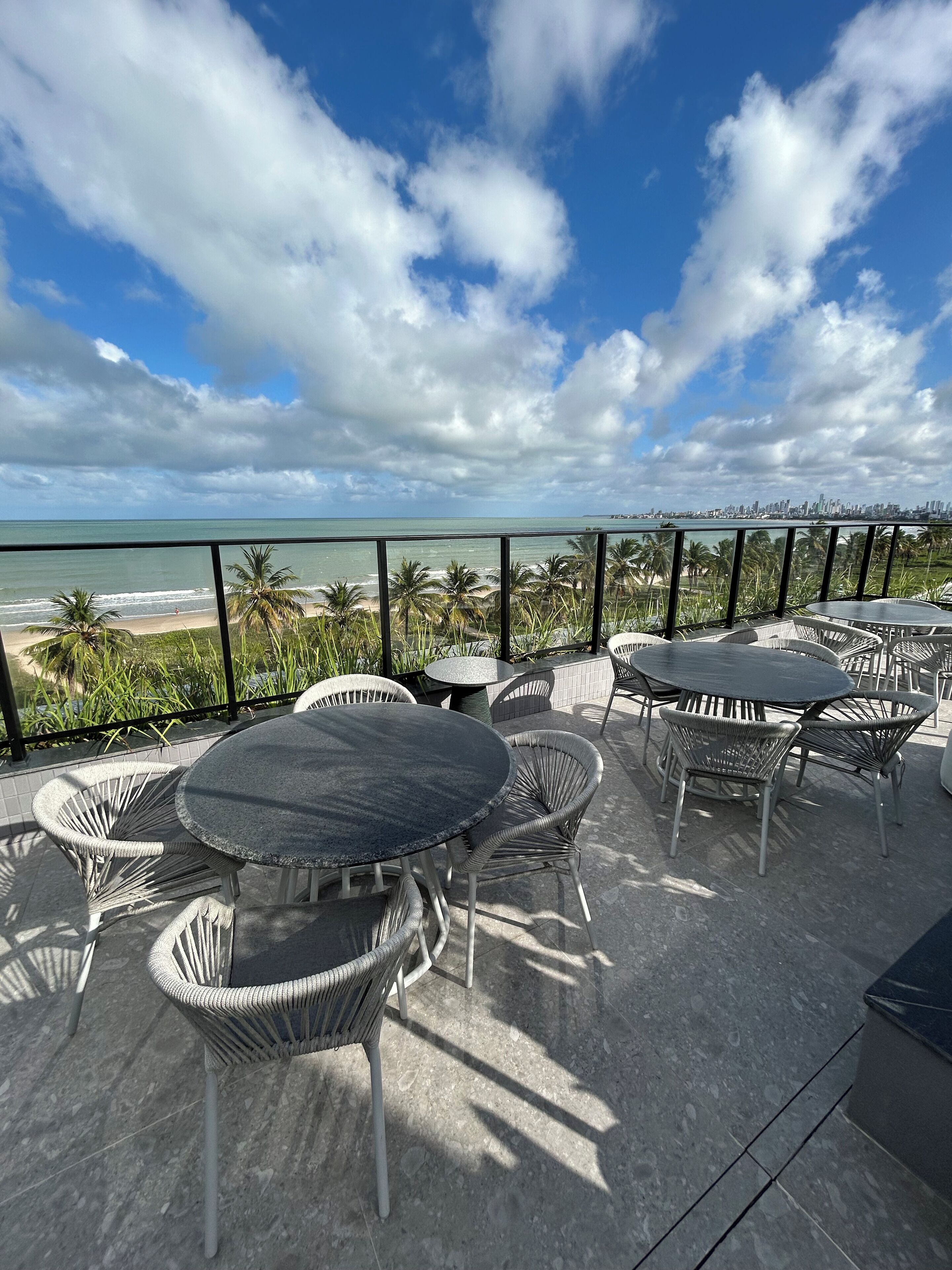 An outdoor patio with round tables and wicker chairs, overlooking a beach and city skyline.