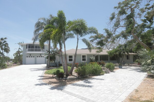 A two-story house with a driveway leading to a garage, surrounded by palm trees and various shrubs.