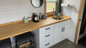 A compact kitchen with a wooden countertop, a sink, a coffee maker, and a mirror.