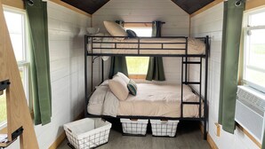 A bunk bed room with a sloped ceiling, wooden walls, and a window with curtains. There are storage bins under the bed.