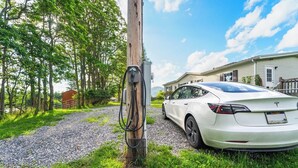 A white Tesla Model 3 parked at a charging station in a rural setting with a house and trees in the background. - 3BR Home Near Snowshoe | Tesla | Pet Friendly  | Elk Mountain Apres’ (Marlinton)