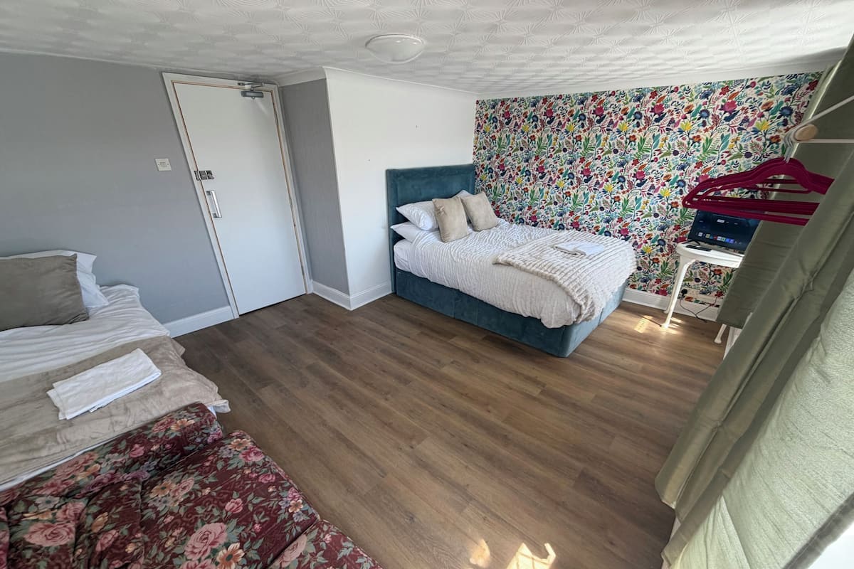 Ein Schlafzimmer mit einem Bett, einem Schlafsofa, einem Schreibtisch mit Computer und einer Wand mit bunten Blumenmustern.