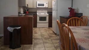 A compact kitchen with a microwave, stove, and refrigerator. There is a small trash bin and a dining area with chairs. - Downtown Hideaway (Fort McMurray)