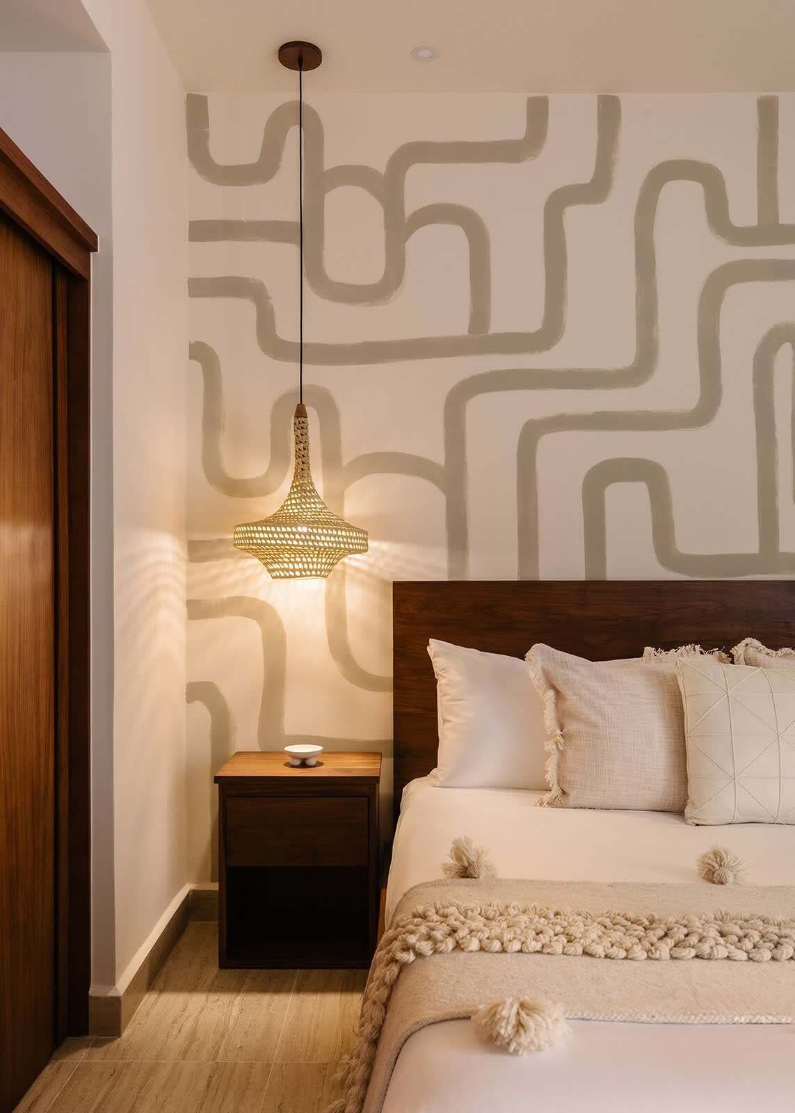 A bedroom with a patterned wall, a hanging light fixture, a wooden nightstand, a bed with multiple pillows, and a knitted throw blanket.