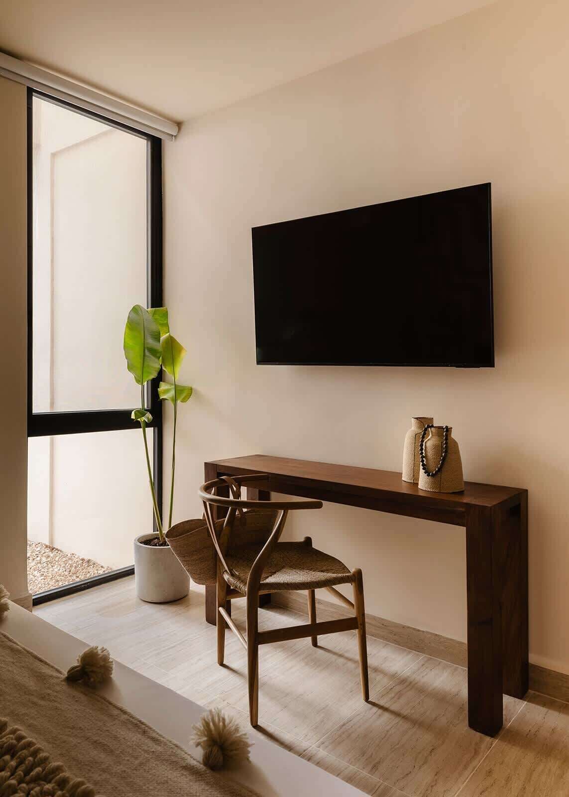 A modern living room with a large window, a flat-screen TV mounted on the wall, a wooden bench, a woven chair, and a potted plant.