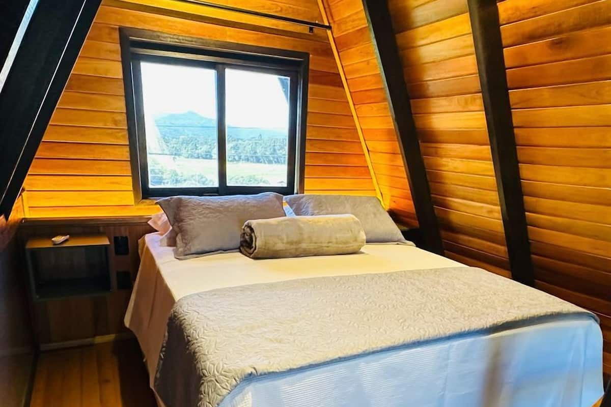 Un dormitorio acogedor en el ático, con techo inclinado de madera, cama con ropa de cama blanca, ventana con vistas al exterior y una pequeña mesita de noche.