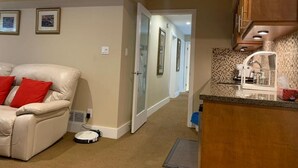 En stue med en beige sofa, en robotstøvsuger og et køkkenområde med bordplade og vask.