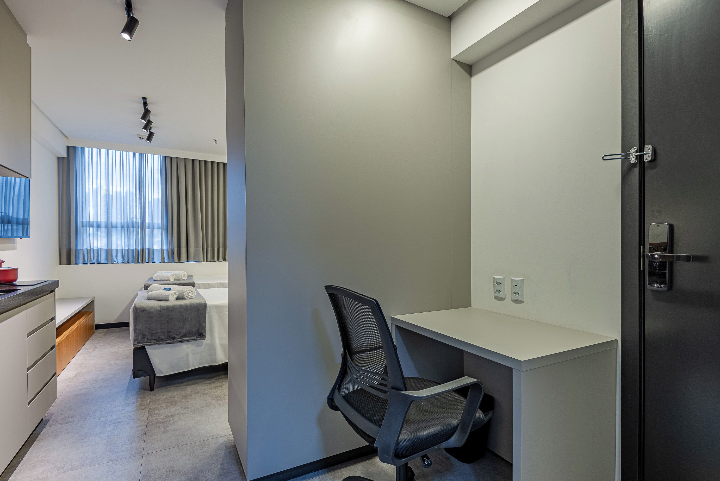 A modern hotel room with a bed, a desk, and a chair. There is a window with curtains, a door, and a light fixture on the ceiling.