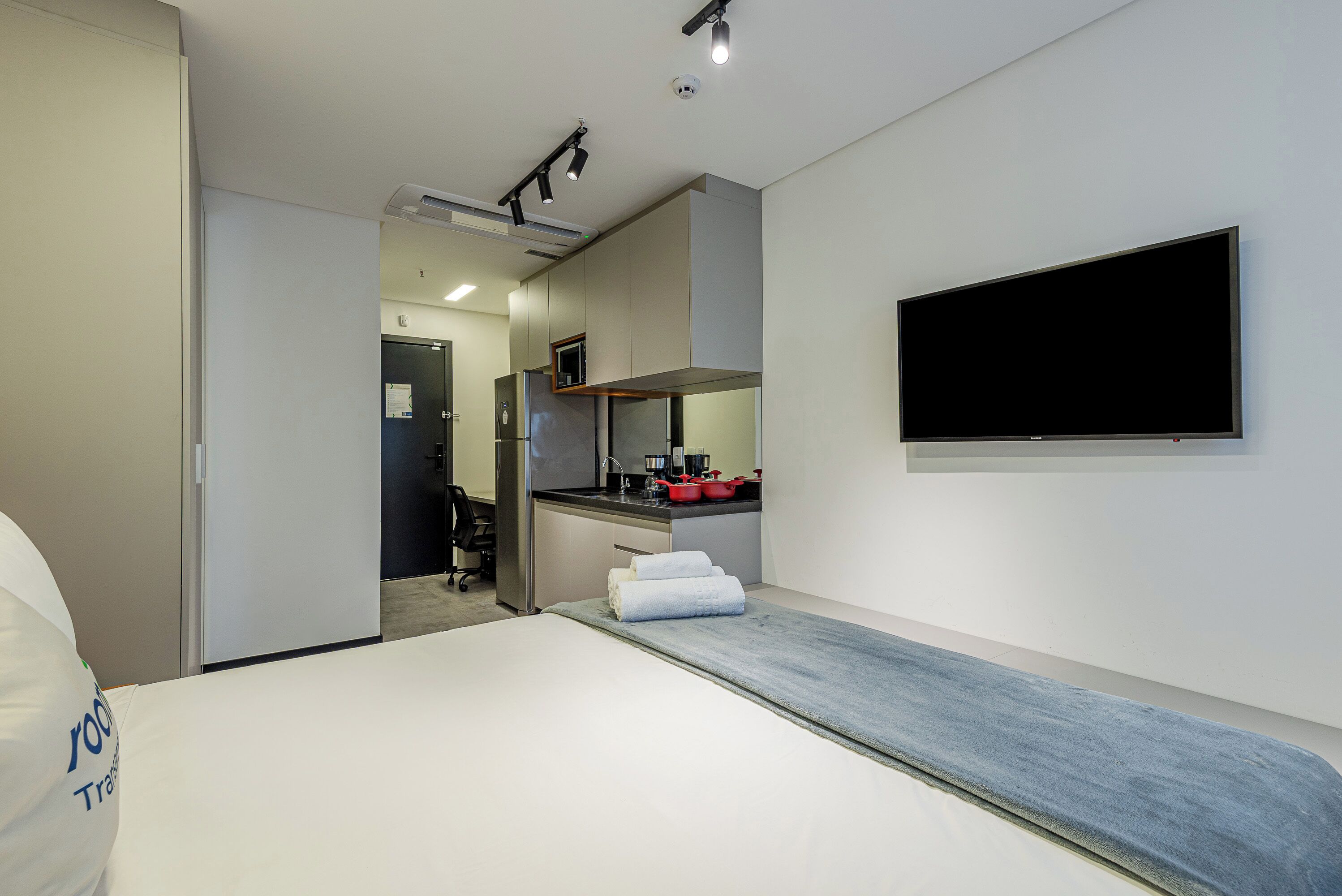 A modern hotel room with a bed, a TV mounted on the wall, a kitchenette, and a bathroom visible through an open door.
