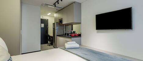 A modern hotel room with a bed, a TV mounted on the wall, a kitchenette, and a bathroom visible through an open door.