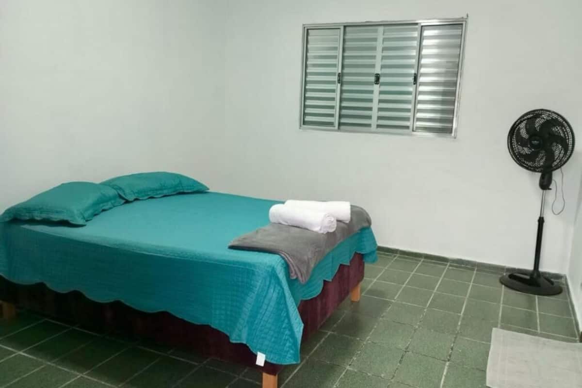 A single bed with a teal bedspread, a window with blinds, and a standing fan.
