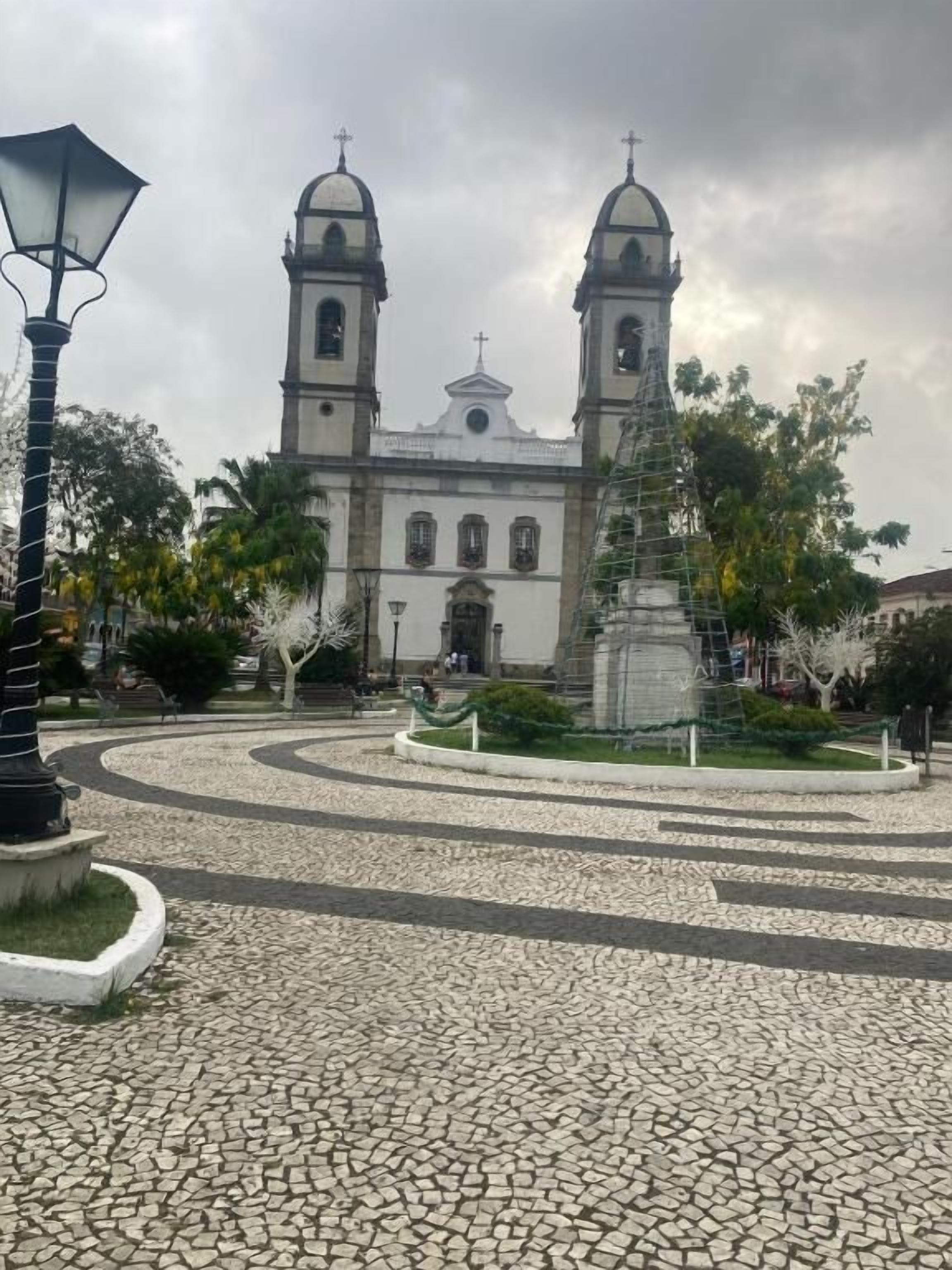 Una iglesia con dos torres y una cúpula central, rodeada de una plaza adoquinada con una estatua y un farol.