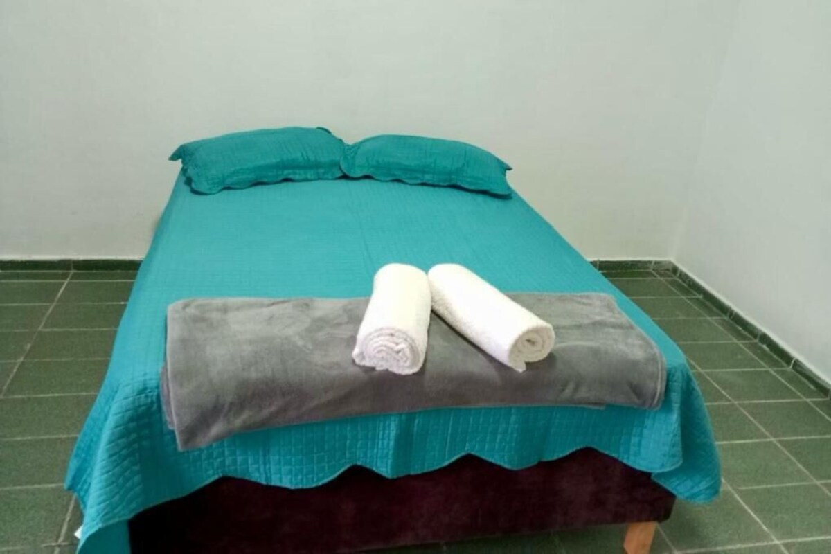 A bed with teal bedding, two rolled white towels, and a grey blanket.