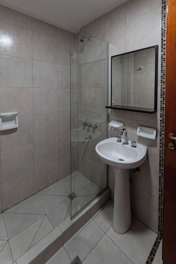 Un baño con cabina de ducha de vidrio, lavamanos pedestal y espejo sobre él.