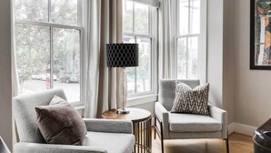A room with two armchairs, a small round table, and a floor lamp. Large windows with curtains are visible, offering a view of the street outside. - Bright & Modern 3BR | Walk to Dining and Park (Richmond)