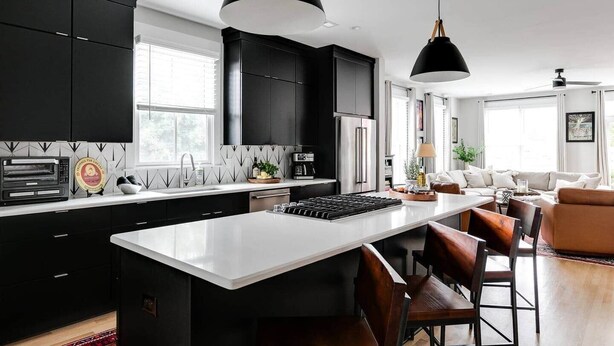 A modern kitchen with black cabinets, a white countertop, and a central island with bar stools. There is a dining area with a sofa and a coffee table. - Bright & Modern 3BR | Walk to Dining and Park (Richmond)