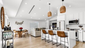 A modern kitchen with white cabinets, a central island with bar stools, a dining area with a wooden table and chairs, and a decorative wall piece.