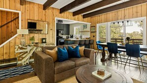 Acogedor interior de cabaña con detalles de madera, sala de estar con sofá y mesa de centro, comedor con sillas y cocina a la vista a través de un umbral abierto.