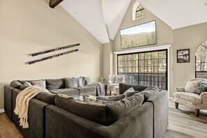 A spacious living room with a grey sectional sofa, a white armchair, and a large window offering a view of trees.