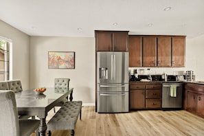 A modern kitchen with a large stainless steel refrigerator, wooden cabinets, and a dining area with a glass table and cushioned chairs.