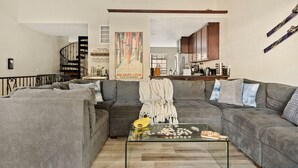 A living room with a gray sectional sofa, a glass coffee table, and a kitchen area in the background. - Edge of the Run @ Snow Summit - Easy Walk to Lifts (Big Bear Lake)
