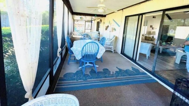 En overdækket terrasse med et bord og stole, en ventilator i loftet og udsigt til et tilstødende rum gennem glasdøre.