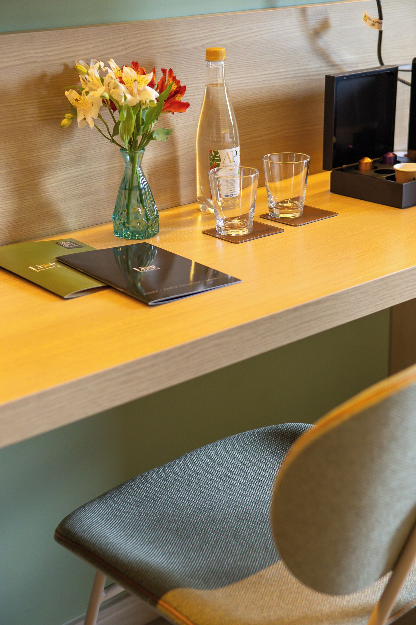 A wooden desk with a vase of flowers, a water bottle, two glasses, and a tablet.