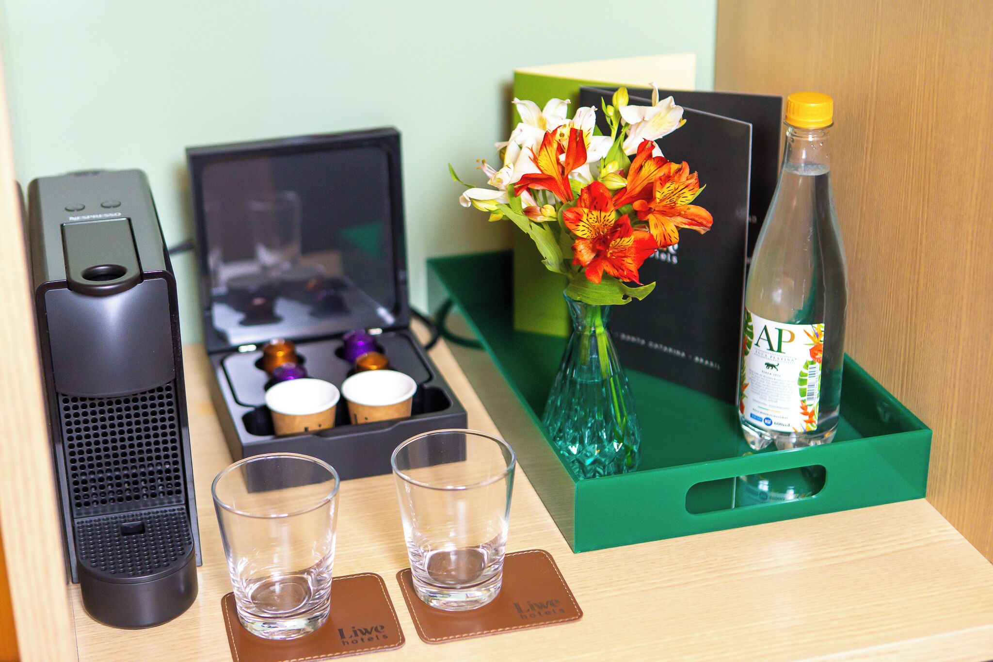 Un mini-bar d’hôtel comprenant une machine à café, deux verres, une bouteille d’eau et un vase avec des fleurs.