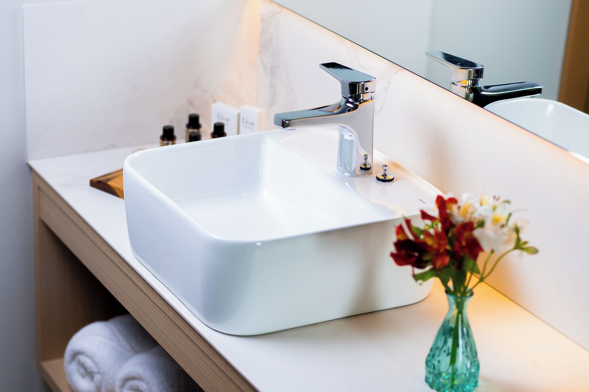 A modern bathroom with a white sink, chrome faucet, and a mirror reflecting the sink. There are toiletries on the countertop and a vase with flowers.