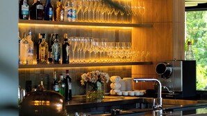 Un área de bar moderna con estantes llenos de botellas de licor y vasos, una máquina de café y sillas de madera.