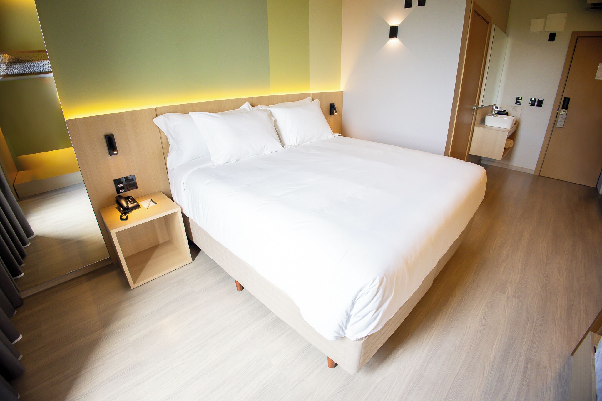 A modern hotel room with a large bed, bedside tables, a small bathroom, and a wooden floor.