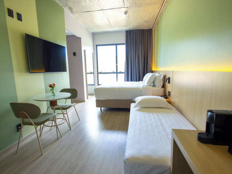 Habitación de hotel con cama, escritorio con cafetera, televisor montado en la pared y ventana con cortinas.