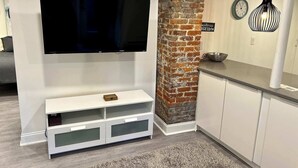 A modern living room with a flat-screen TV mounted on a white wall, a brick accent wall, a white entertainment unit with drawers, and a grey shaggy rug.