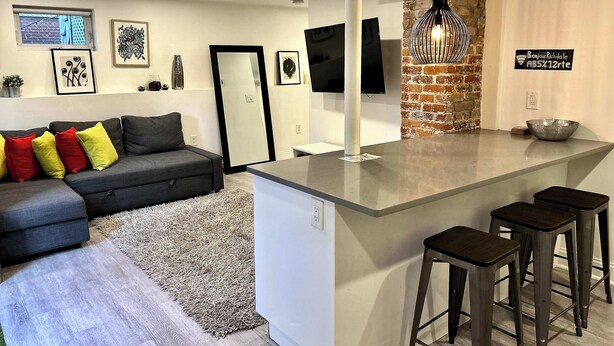 A modern kitchen with a central island, bar stools, a grey sofa with colorful cushions, and a brick accent wall.