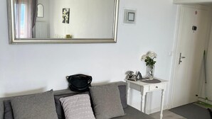 A modern living room with a grey sofa, a large mirror, a small white table with a vase, and a door in the background.