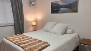 Un dormitorio con cama, lámpara de noche, ventilador de pared, ventana con cortinas y un cuadro en la pared.