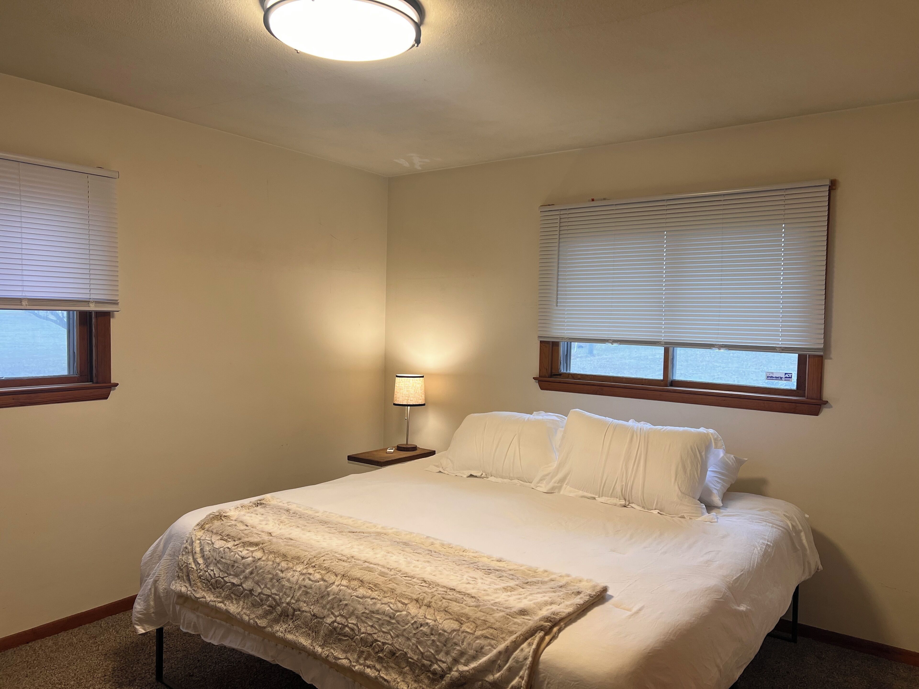 A bedroom with a bed, a nightstand, a lamp, and two windows with blinds.