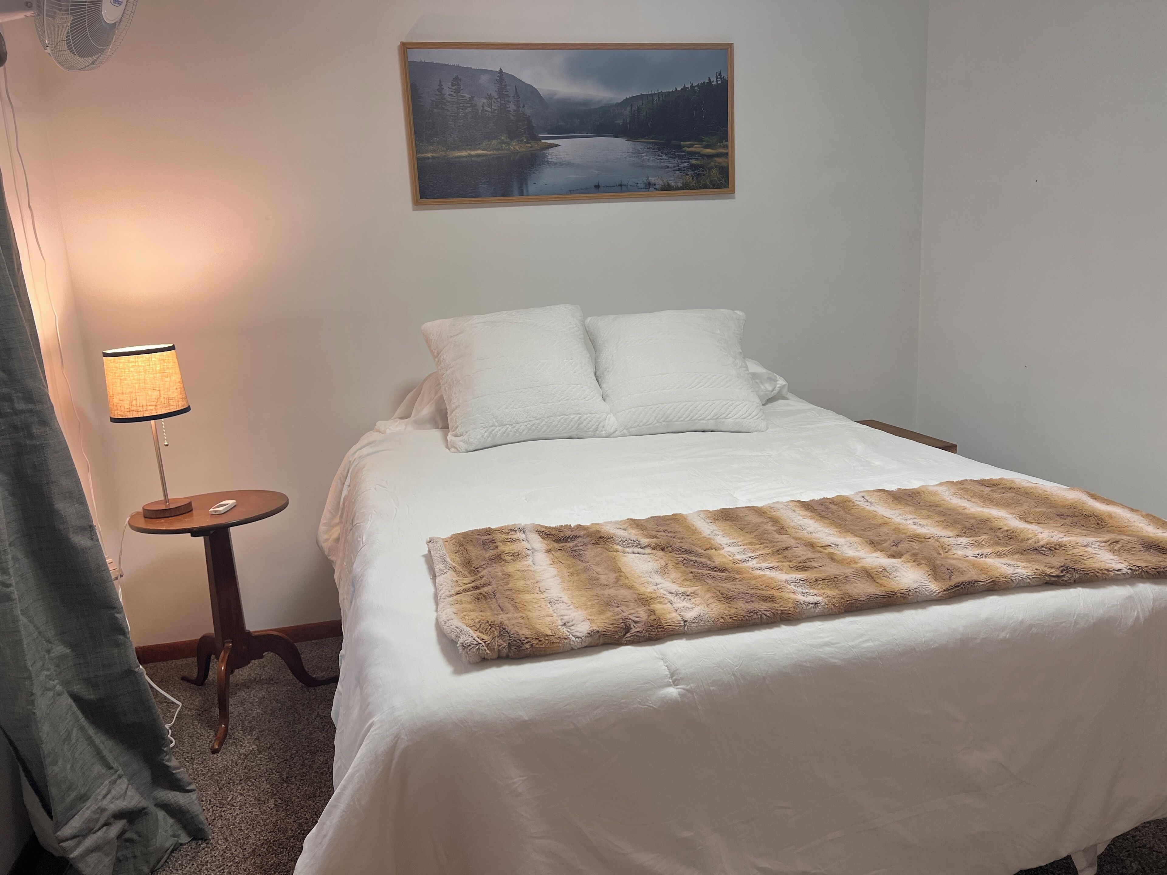 A neatly made bed with white bedding and a brown fur throw, a bedside lamp, and a framed landscape painting above the bed.