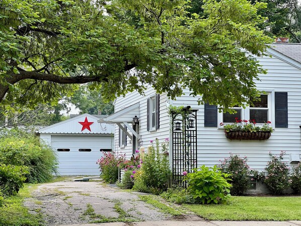 Una casa blanca con una estrella roja en la puerta del garaje, rodeada de follaje y flores.