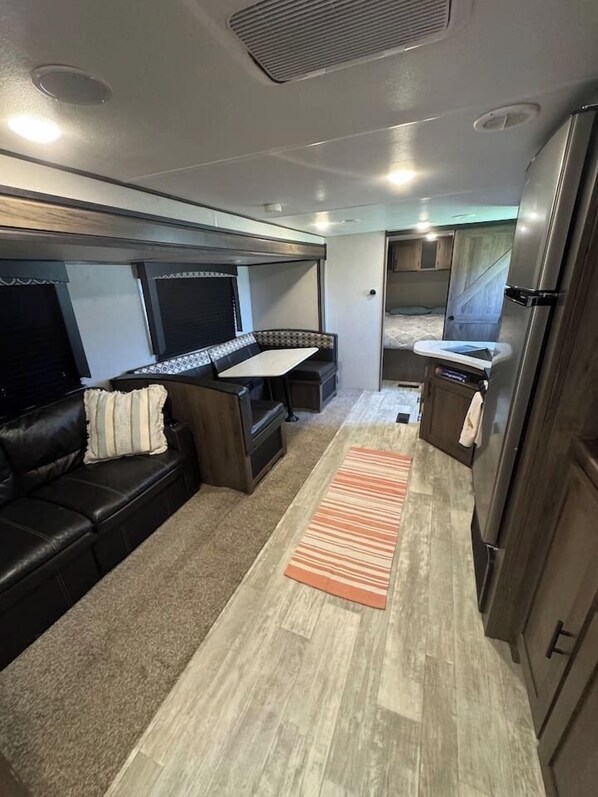 A compact RV interior with a sofa, a small table, a kitchen area with a refrigerator, and a window.