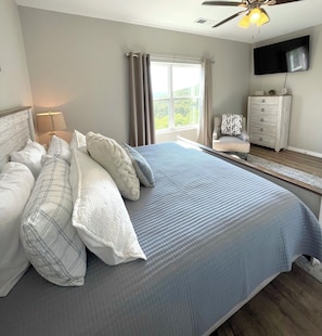 Un dormitorio con una cama grande, una mesita de noche, una lámpara, una silla, un aparador y un televisor en la pared.